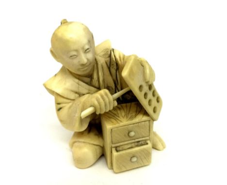 A Meiji Period Ivory Figure:Carved in the form of a seated man holding a holed board and stick and a two drawer chest at his 