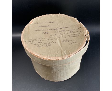 Round cardboard box containing an elaborate baby's bonnet of ivory silk trimmed with lace and ribbons. Written in ink upon th