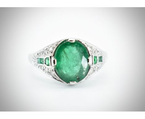 An 18ct white gold emerald and diamond ring. The ring in the Art Deco style being set with a central oval mixed cut emerald h