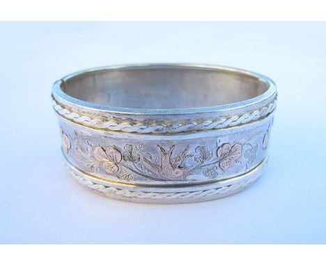 A silver gold stiff hinge bangle depicting swallow in foliage within rope twist border, unmarked, inscription to back