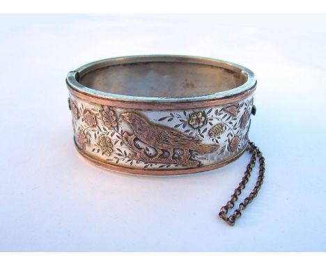 A silver gold stiff hinge bangle with bird in branches with acorns and flowers, with safety chain, unmarked
