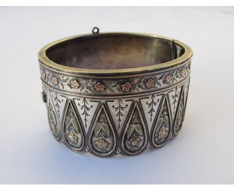 A silver gold stiff hinge bangle, pear drop panels of flowers under a wavy edge unmarked
