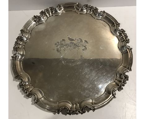 A George V silver salver with foliate scrollwork decorated pie-crust rim, the centre field decorated with an armorial of scro