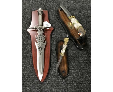 A fantasy knife on wooden shelf together with two hunting knives with animal grips on stands 