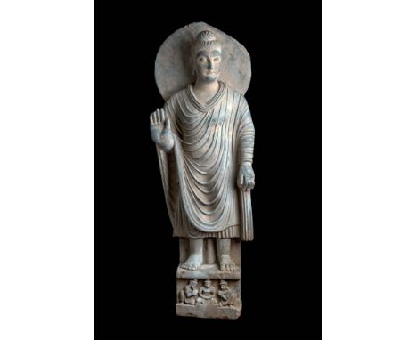 Ca. AD 200 - 300.A grey schist statue of Buddha standing atop an integral rectangular pedestal. The figure's left leg is slig