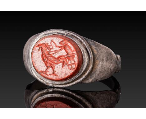 Ca. AD 1 - 300. An oval red jasper intaglio engraved with a fantastical scene of a rooster advancing to the left, harnessed t