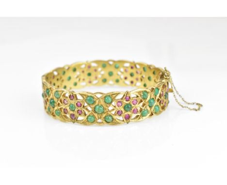 An Indian gold plated, ruby and emerald set bangle, with pierced floral fretwork design inset with round cut rubies and caboc