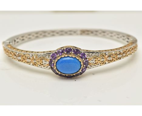 A WHITE METAL HINGED BANGLE, set with a blue cabochon stone within a surround of twelve oval cut amethysts, yellow metal scro