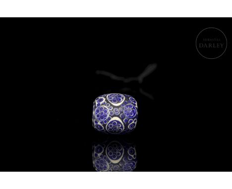 A vitreous-paste bead with blue and white enamel. It is oval, pierced by a large central hole, and decorated with small circl