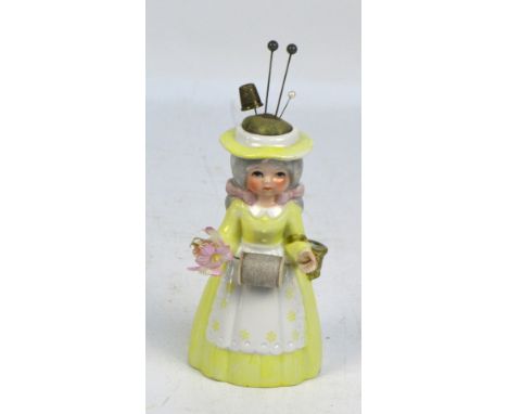 A ceramic pedlar doll sewing companion, with pin cushion hat, reel suspended between hands and a small thimble in her basket,