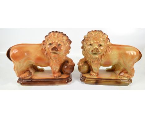 An opposing pair of 20th century ceramic lions with glass inset eyes, each with front paw leaning on a ball, on rectangular b