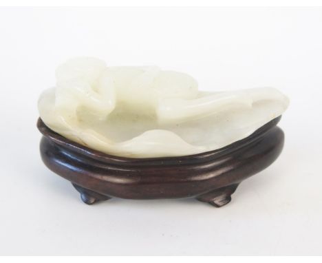 A Chinese carved jade figure of a reclining female figure, lying on a leaf, on a polished wood stand, 7cm wide. 