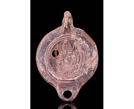 Ca. 100-200 AD A figural terracotta oil lamp with a single handle. The discus is framed by two concentric rings, in the middl