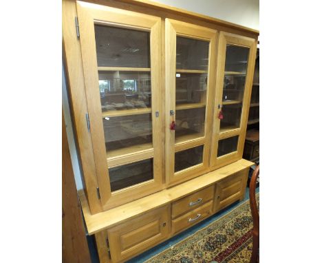 A light oak wall unit display cabinet with three glazed doors enclosing shelves above two short drawers flanked by cupboard d