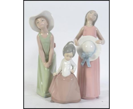 Two Lladro girls one hatted and leaning forward wearing a green dress.  The other is standing upright wearing a pink dress an