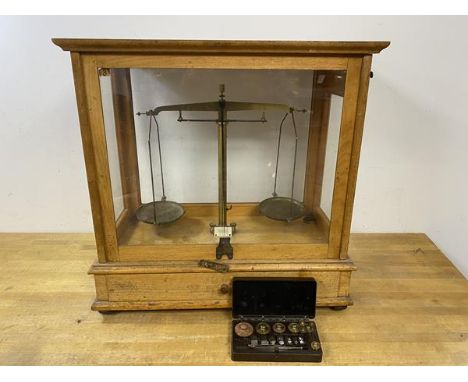 An early 20thc set of scales in wooden and glazed showcase, with fitted drawer to base, back foot lacking, and boxed set of w