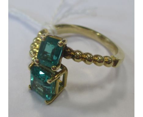 A two stone emerald ring, the two square step cut emeralds, each in a four claw setting, set to either end of a slightly over