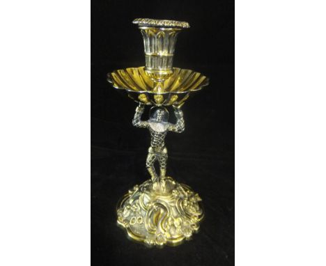 A William IV silver gilt taper stick stand in the form of a harlequin holding a sconce, standing on a floral embossed base, 1