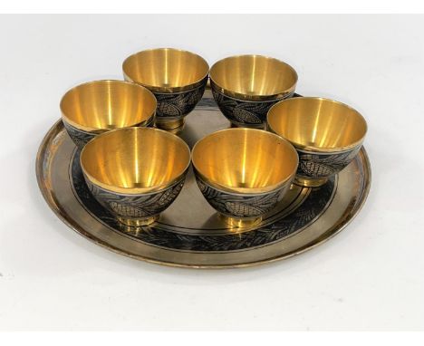 A Russian white metal set of six vodka cups and a circular tray decorated in gilt in the Niello manor, stamped to base with s