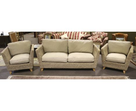 A contemporary rattan three-piece sofa suite. Including two armchairs and a sofa, with cream upholstered cushions, on taperin