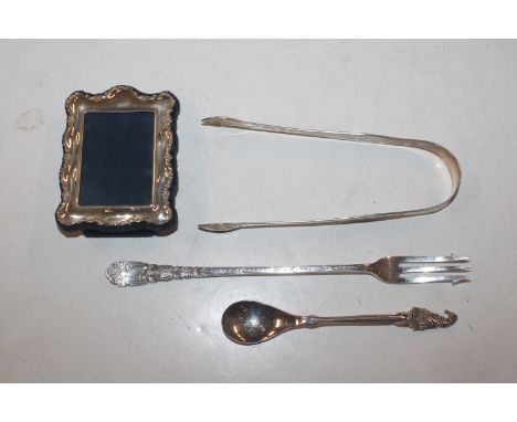 A small silver mounted photo frame; a pair of silver bright cut sugar tongs; a silver pickle fork and a white metal spoon