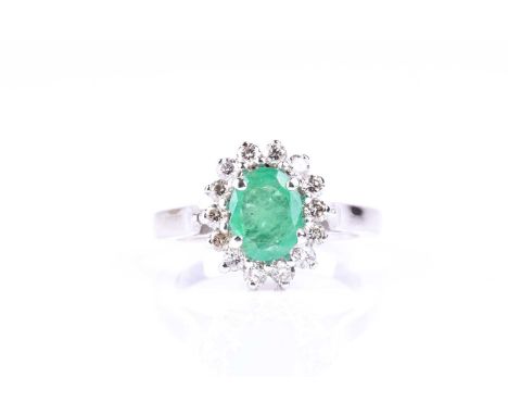 An emerald and diamond cluster ring, the oval cut emerald, in four claw mount within a border of briliant cut diamonds to pla