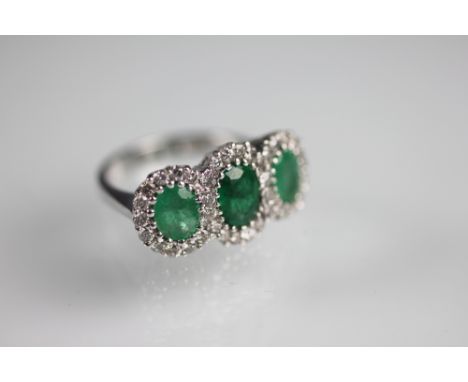 An emerald and diamond triple cluster ring, the three oval, graduated emeralds within a diamond set surround, all set in whit