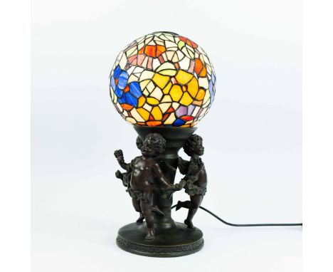 A TIFFANY STYLE TABLE LAMP. the stained glass leaded shade of globular form, raised on three cherubs in bronze finish, circul