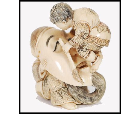 A Japanese Meiji period carved ivory netsuke in the form of a theatrical mask raised by a seated figure with further figure a