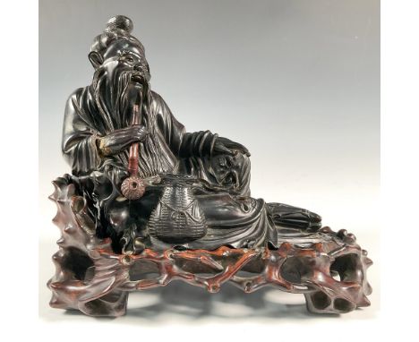Intricately carved seated figure of Luohan fisherman in reclining position with a pipe in hand. Surmounted on a dark carved w