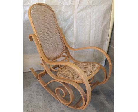 A light wood rocking chair with cane seat and back. Scroll detail above rockers. 