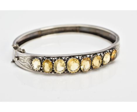 A WHITE METAL CITRINE HINGED BANGLE, designed with a row of graduated claw set, oval cut citrines, openwork and applied bead 