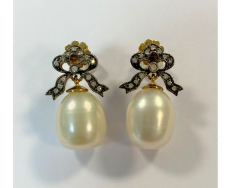 each with a decorative bow set with small round brilliant cut diamonds and suspending a 12mm long oval pearl of a pale creamy