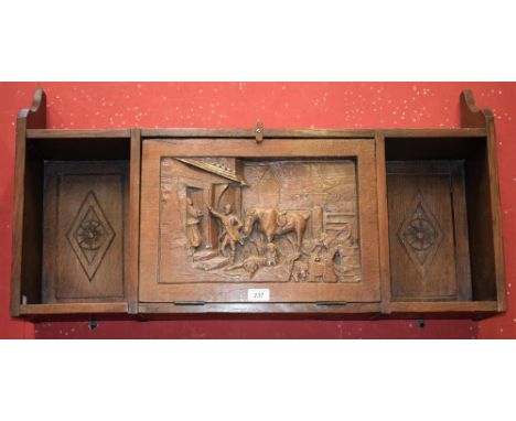 A carved oak wall hanging display cabinet shelf, central carved fall front decorated with a farm and stable yard, between ope