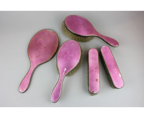 A George V five piece silver and pink enamel backed dressing table set comprising a hand mirror, two hair brushes and two clo