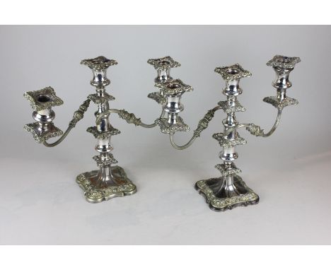 A pair of silver plated three-light candlesticks, with ornate, foliate and scrolled borders, on baluster stems and square bas