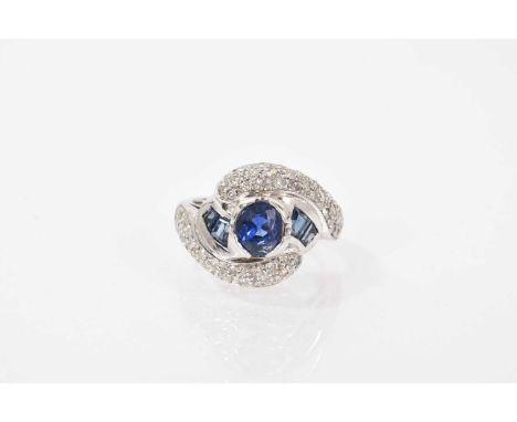 Art Deco style sapphire and diamond cross over ring with a central oval mixed cut blue sapphire flanked by calibre cut sapphi