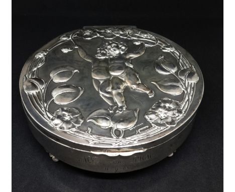 An Edwardian round silver trinket box the hinged lid depicting two cherubs within a flower and scrolled border, the base with