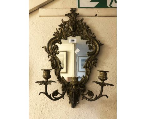 An old cast brass framed ornate wall mirror with masks to top and base, shaped plate and triple candle sconces - one nozzle m