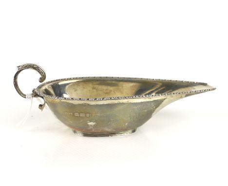 An Art Deco silver shallow oval sauce boat. With a bead and reel rim, a leaf-capped scroll handle and a reeded oval foot, hal