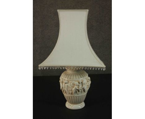 An ivory coloured resin table lamp, cast with a Bacchanalian scene, with a shade. H.67 W.40cm. 