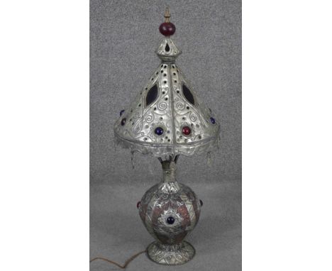 An Indian or Middle Eastern metalware lamp and shade, with embossed designs, glass panels and glass cabochons, the conical sh