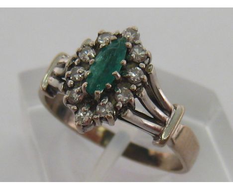 An emerald and diamond cluster ring, the central navette cut emerald 6.8mm long, in a surround of eight cuts to trifurcated s