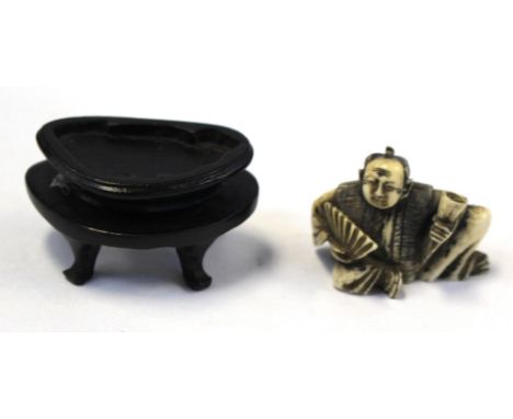 A Japanese carved ivory netsuke, in the form of a seated man with fan and scroll, on stand