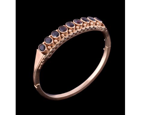  A late Victorian garnet hinged gold bangle,   circa 1900, the oval cut garnets within millegrained collet settings, with bea