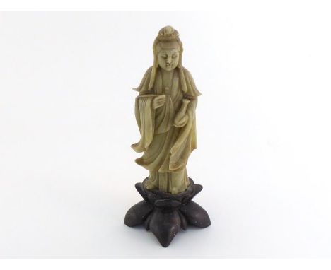 A Chinese carved soapstone figure of Guanyin standing on a lotus shaped soapstone base, wearing robes and holding a vase on l