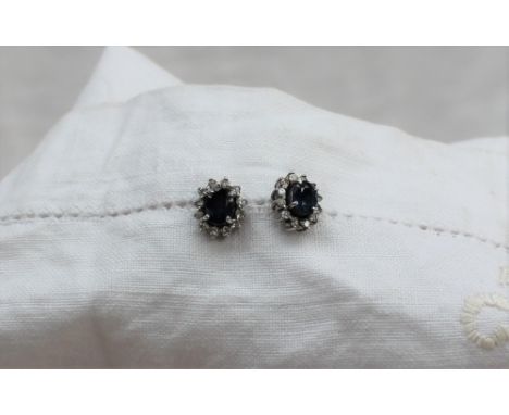 A pair of sapphire and diamond stud earrings, each set with a central oval sapphire approximately 6mm x 5mm, surrounded by tw