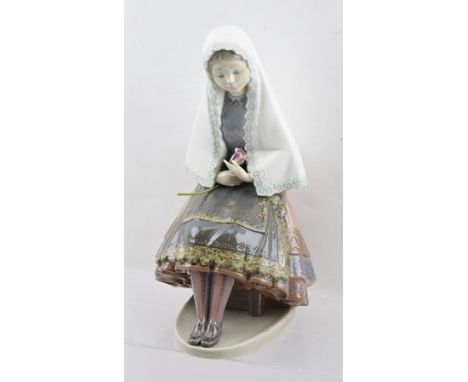A LLADRO FIGURINE OF A YOUNG GIRL in traditional dress with white shawl, a rose in her hand, sitting on a stool, pastel polyc