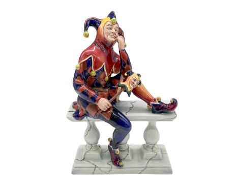 A rare porcelain figure of a court fool seated on a railing and holding a marotte. Red, blue, yellow, and green colorations. 