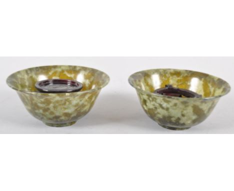 A pair of spinach jade bowls, each of flared form in mottled green hues, with a pair of carved wooden stands, in presentation